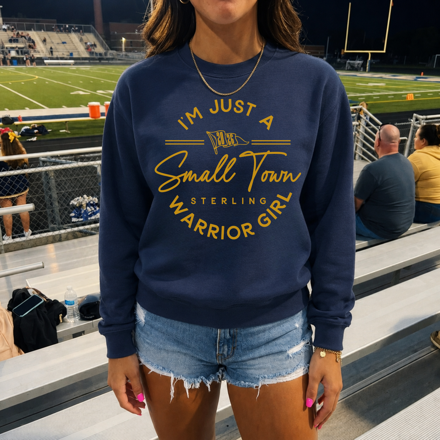 Woman wearing a navy Small Town Warrior Girl Sterling Warriors premium crewneck sweatshirt with denim shorts while watching a football game from the stadium bleachers.