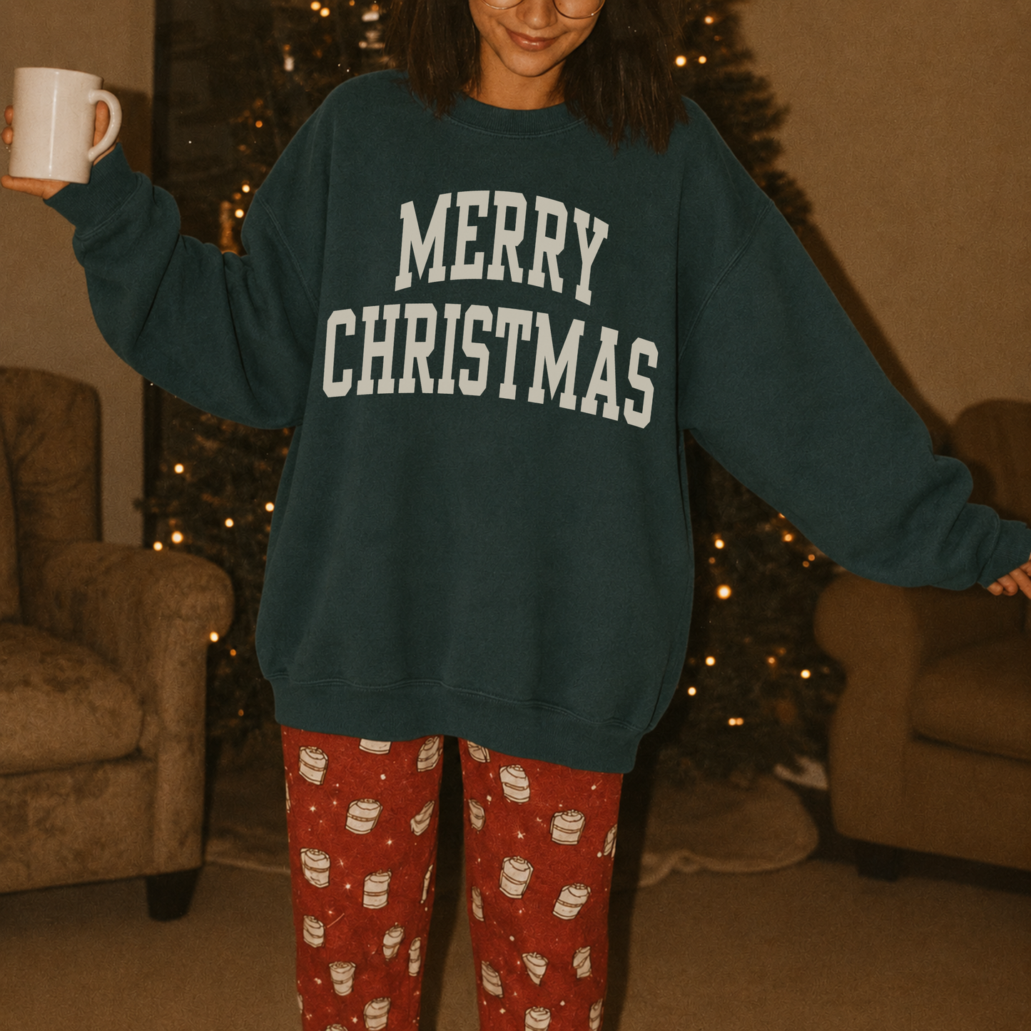 Woman wearing a dark green Merry Christmas varsity sweatshirt with festive Christmas pajama pants while holding a coffee mug beside a Christmas tree.
