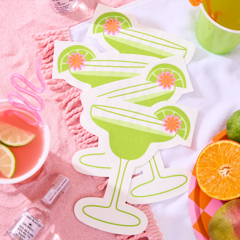 green margarita glass  napkin with lime and strawberry on a pink blanket with drinks and fruit.