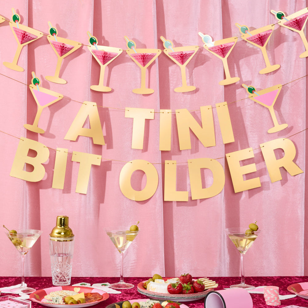 Decorative banner with 'TINI BIT OLDER' text and cocktail-themed decorations on a pink background.