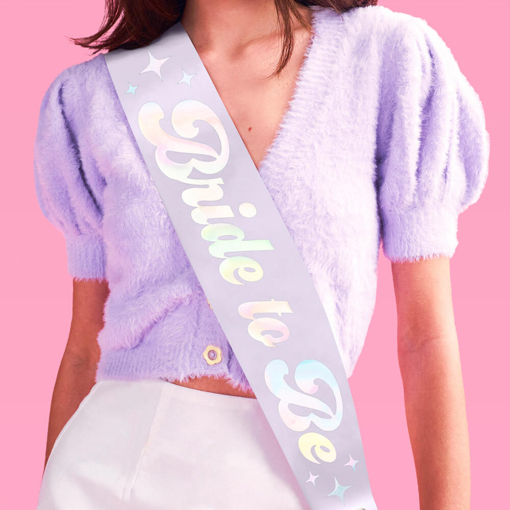 Woman wearing a lavender sweater with a 'Bride to Be' sash on a pink background