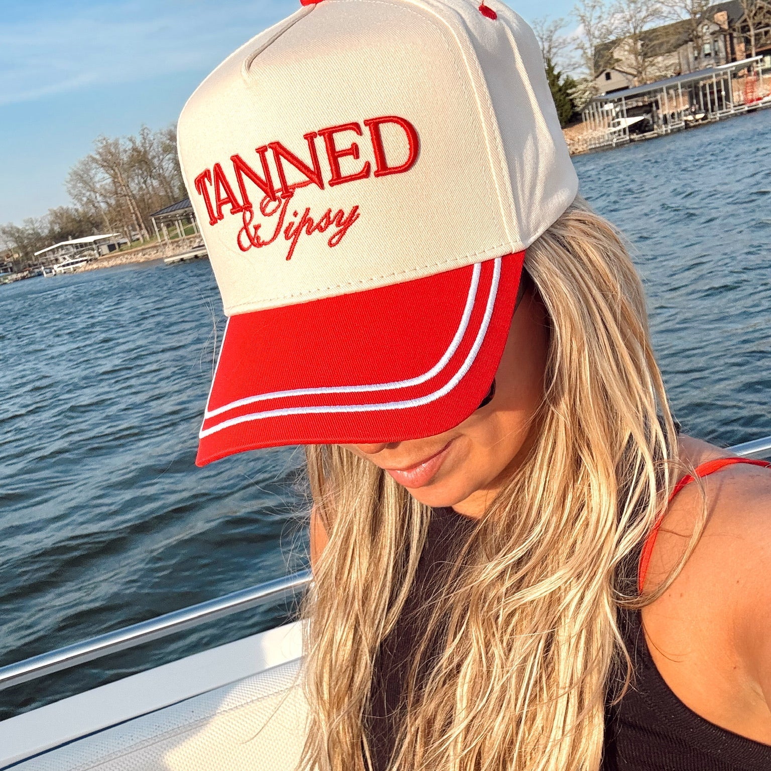 Woman wearing a cap with 'Tanned & Tipsy' text on a boat by a body of water.