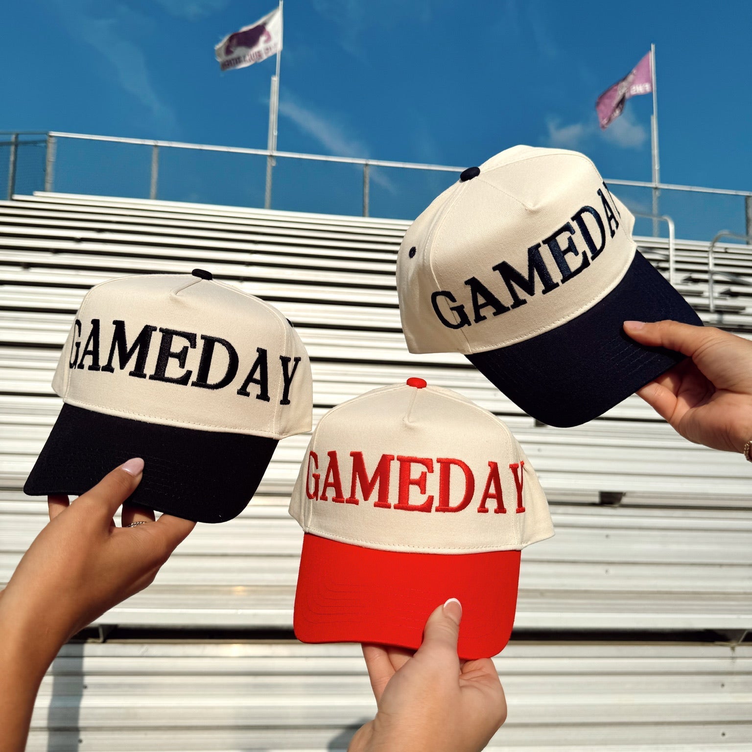 Three 'GAMEDAY' hats held by hands against a stadium background with flags.