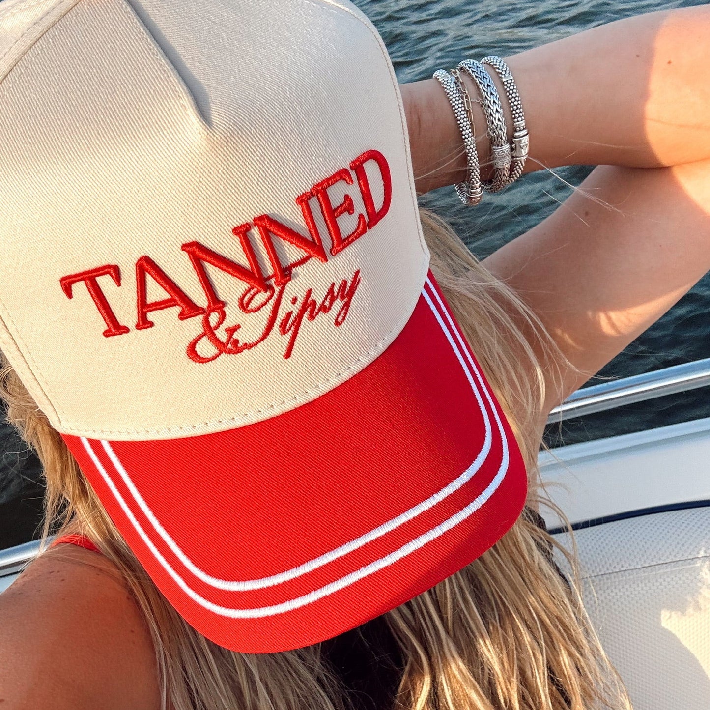Person wearing a red and white cap with 'Tanned & Tipsy' text on a boat.