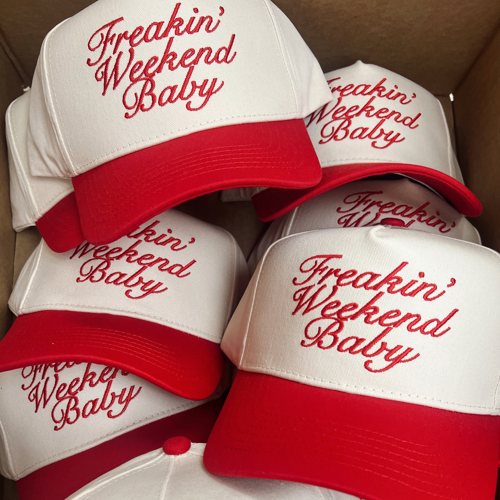 White baseball caps with red brims and 'Freakin' Weekend Baby' text in red, stacked in a box.