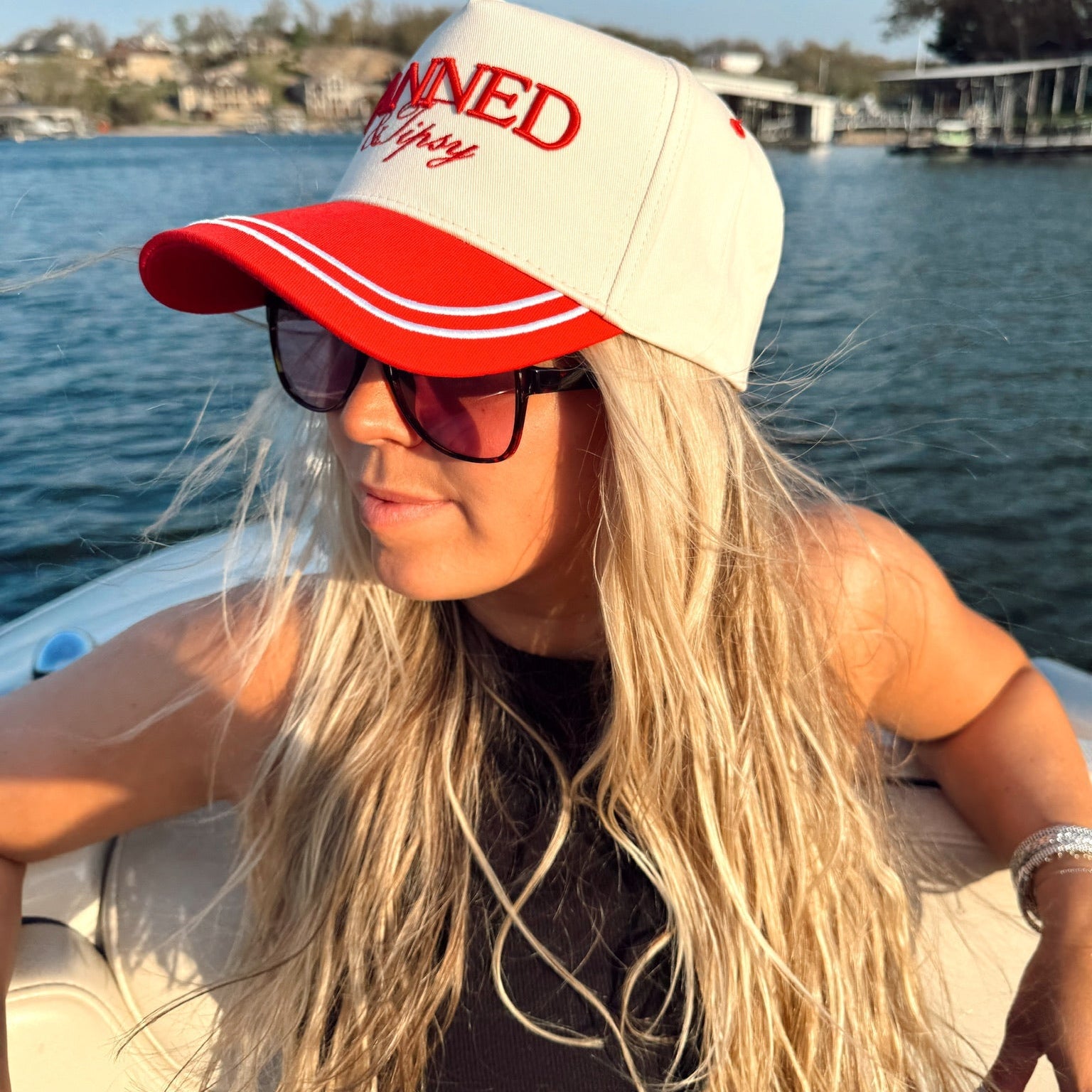 Woman wearing a red and white cap with text, sunglasses, and a black top on a boat with water and sky in the background.