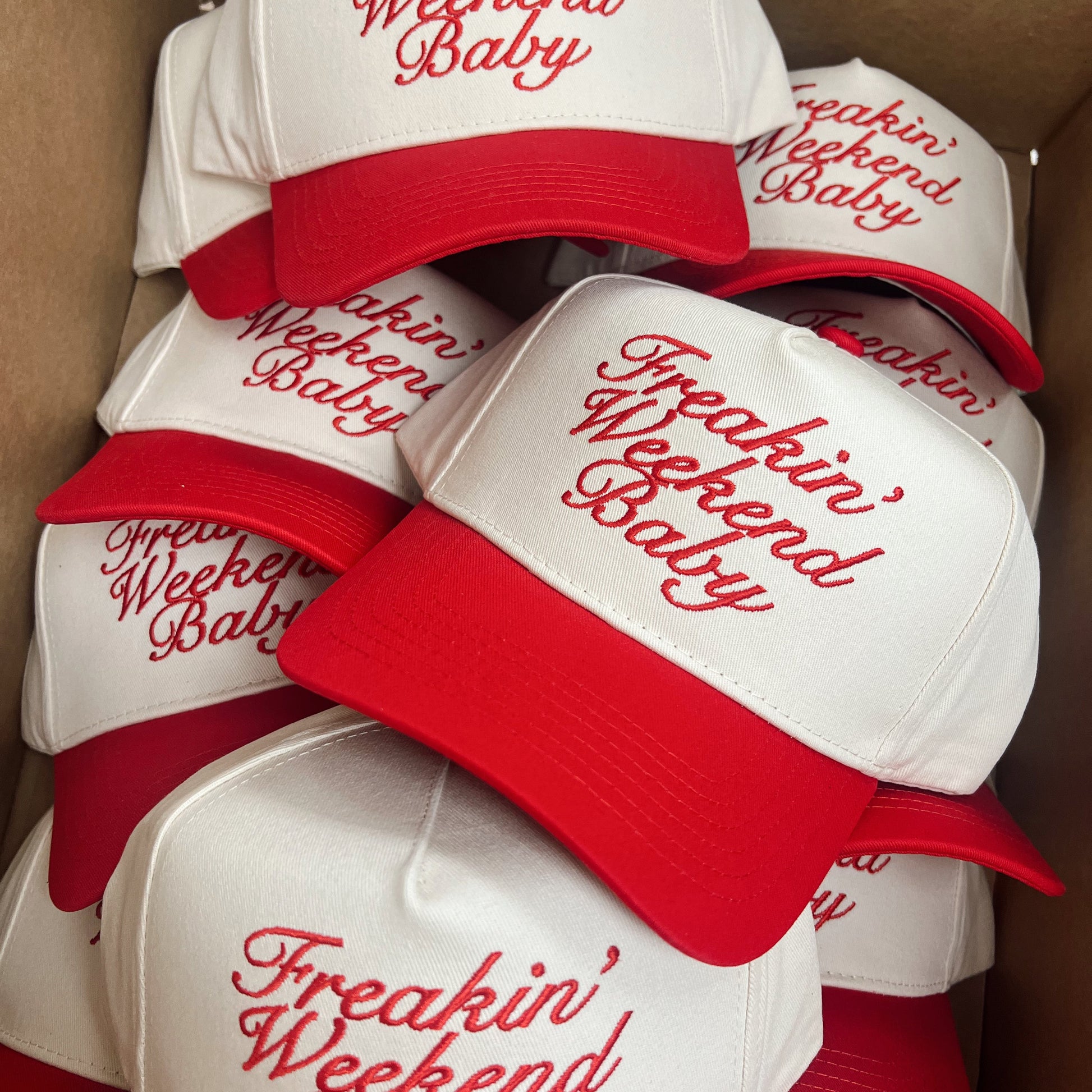 White baseball caps with red brims and 'Freakin' Weekend Baby' text in a cardboard box.
