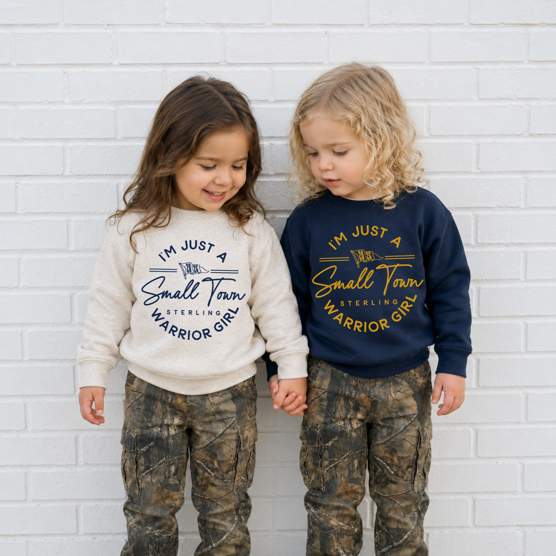 Two toddler girls wearing matching Sterling Warrior Girl football sweatshirts in natural heather and navy with camo pants.