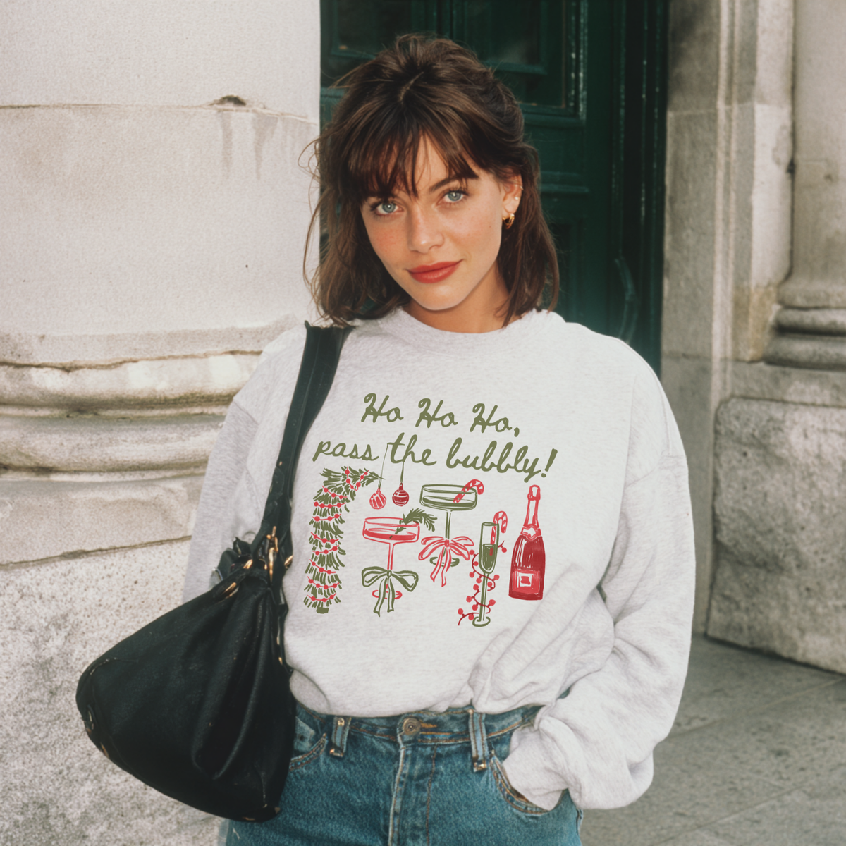 Woman wearing ash grey “Ho Ho Ho, Pass the Bubbly!” sweatshirt with red and green champagne design, standing outside a stone building.