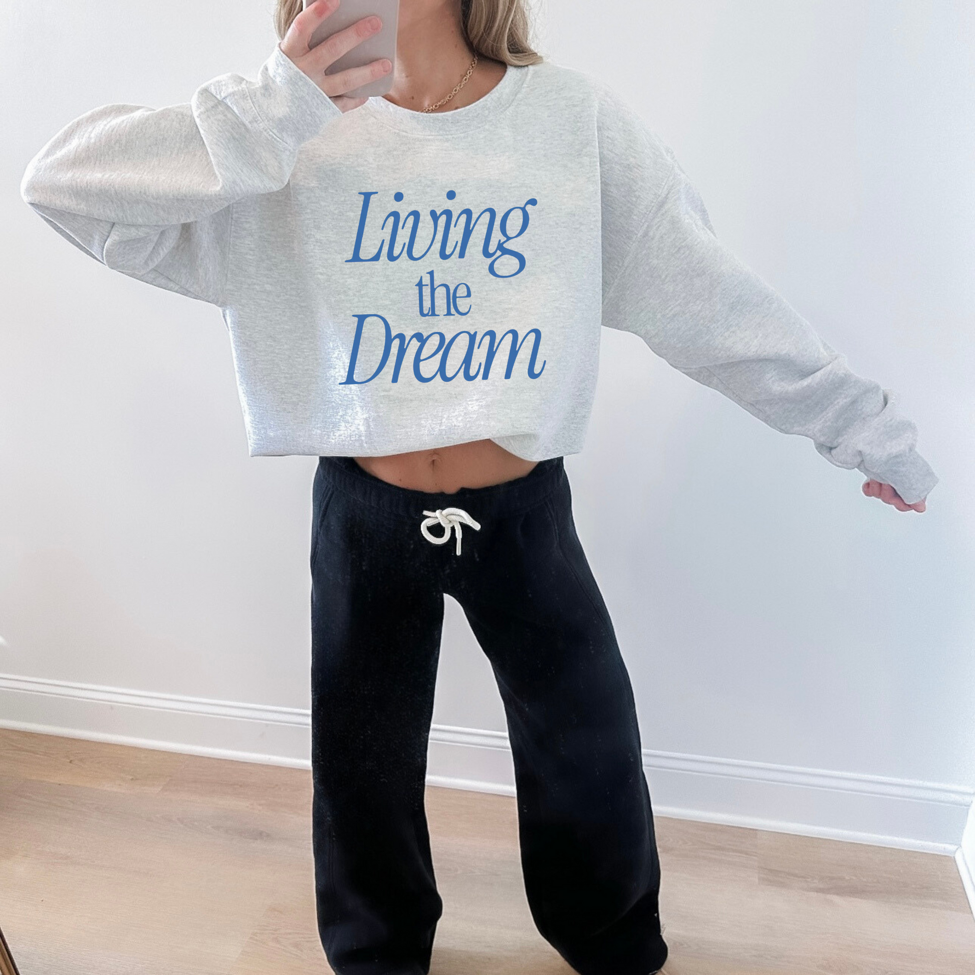 Person wearing a gray sweatshirt with 'Living the Dream' text and black pants, standing against a white wall.