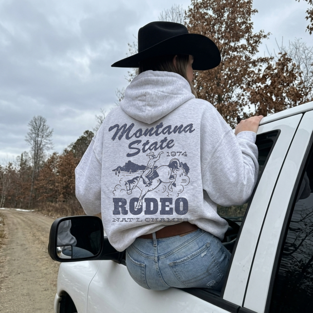 Woman wearing ash grey Montana State Rodeo hoodie with oversized vintage cowboy and bucking horse back graphic beside white truck.