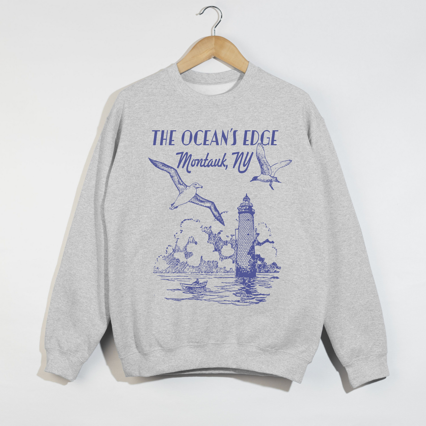 "Ash grey sweatshirt with blue Montauk lighthouse and seagulls coastal design displayed on hanger."
