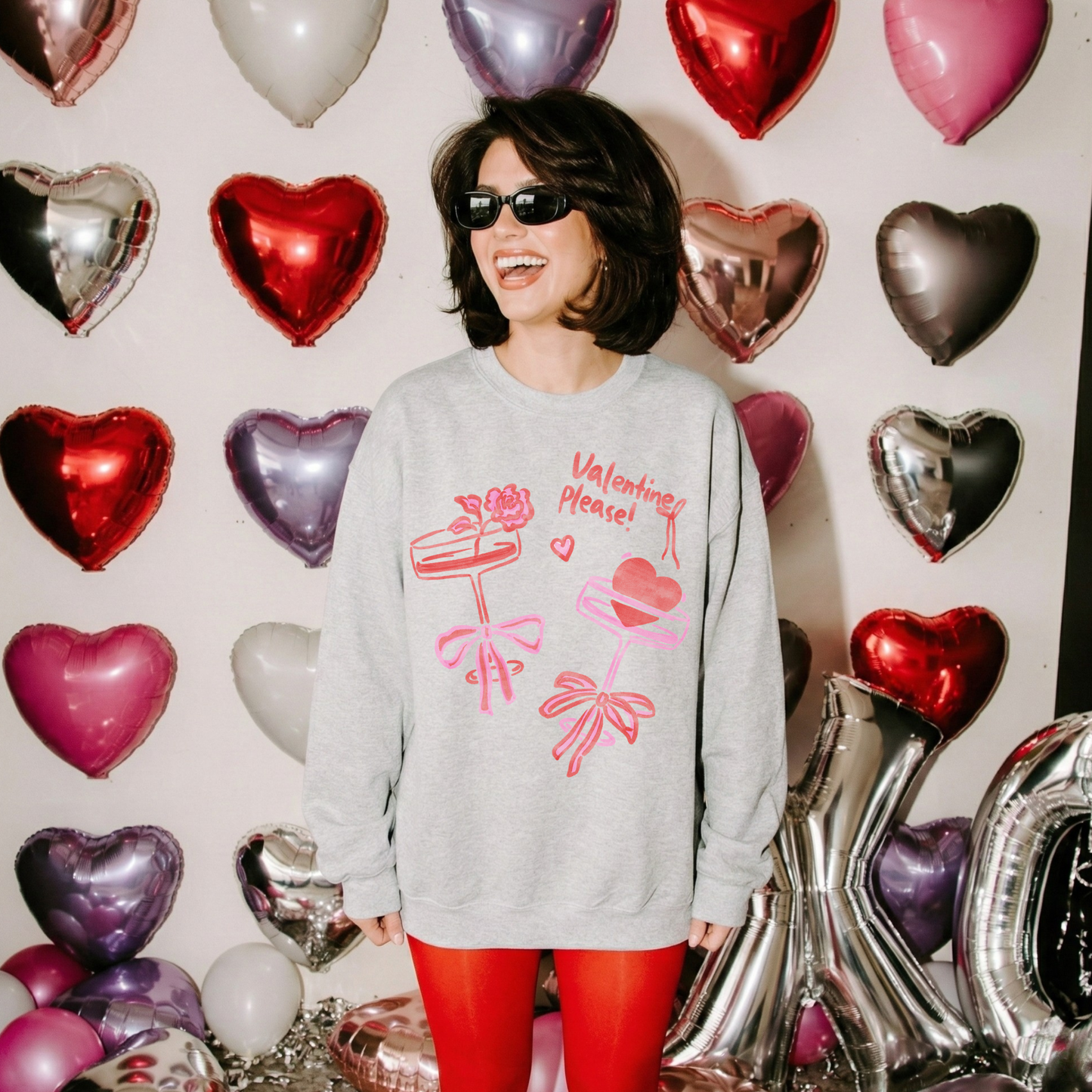 Woman wearing an ash grey Valentine’s crewneck with pink and red bow graphics, laughing in front of a wall of metallic heart balloons.