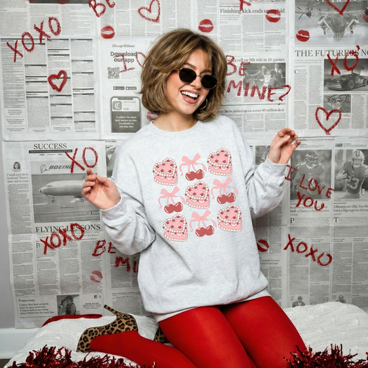Woman wearing an ash grey Valentine sweatshirt with pink cake and cherry graphics, sitting in front of a lipstick-decorated newspaper backdrop.