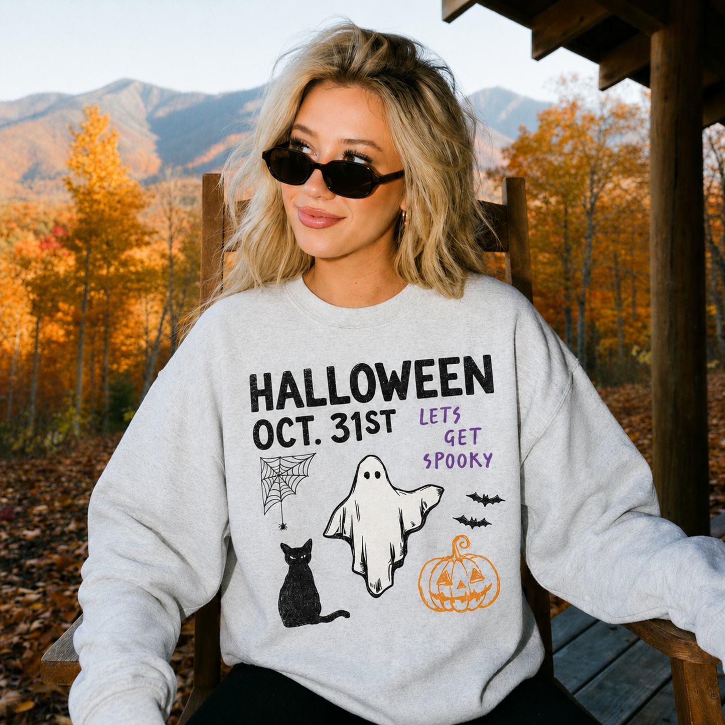 Halloween Oct. 31st ghost graphic sweatshirt featuring a black cat, pumpkin, spiderweb and bats worn outdoors during fall with colorful autumn mountains in the background.