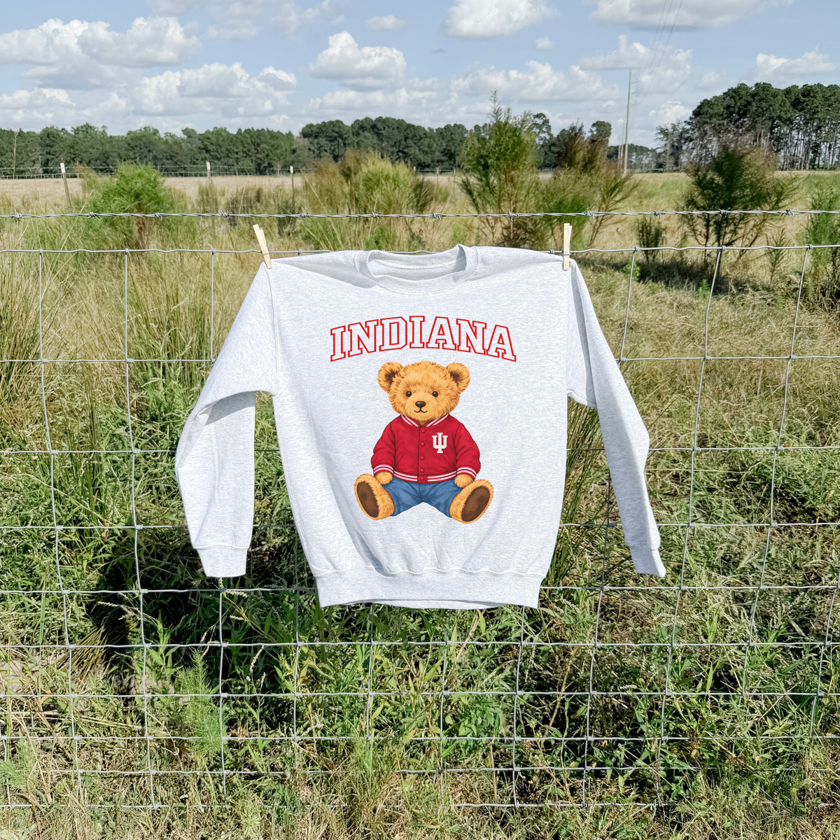 Ash grey Indiana teddy bear sweatshirt hanging on a rustic wire fence outdoors, featuring red arched Indiana lettering and a golden teddy bear wearing a crimson varsity jacket with IU logo.