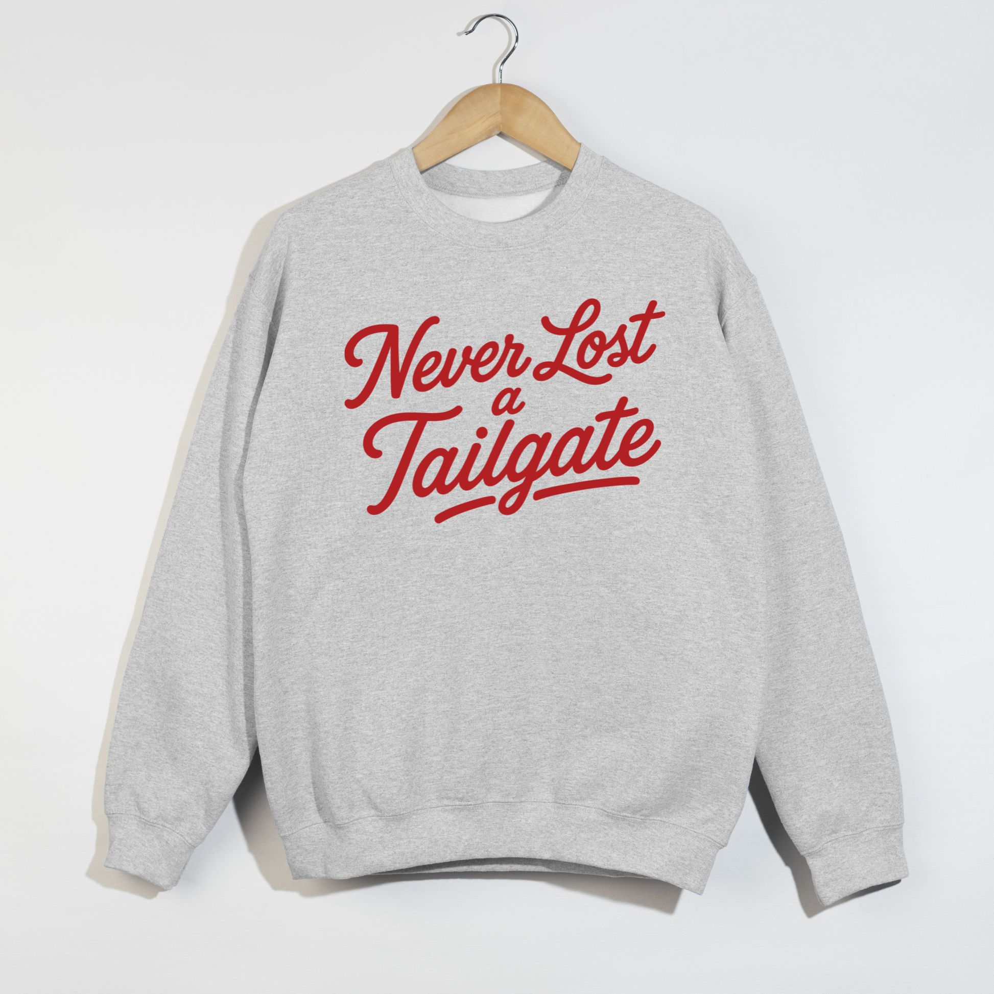 Ash gray sweatshirt featuring the phrase "Never Lost a Tailgate" in bold crimson script lettering hanging on a wooden hanger.