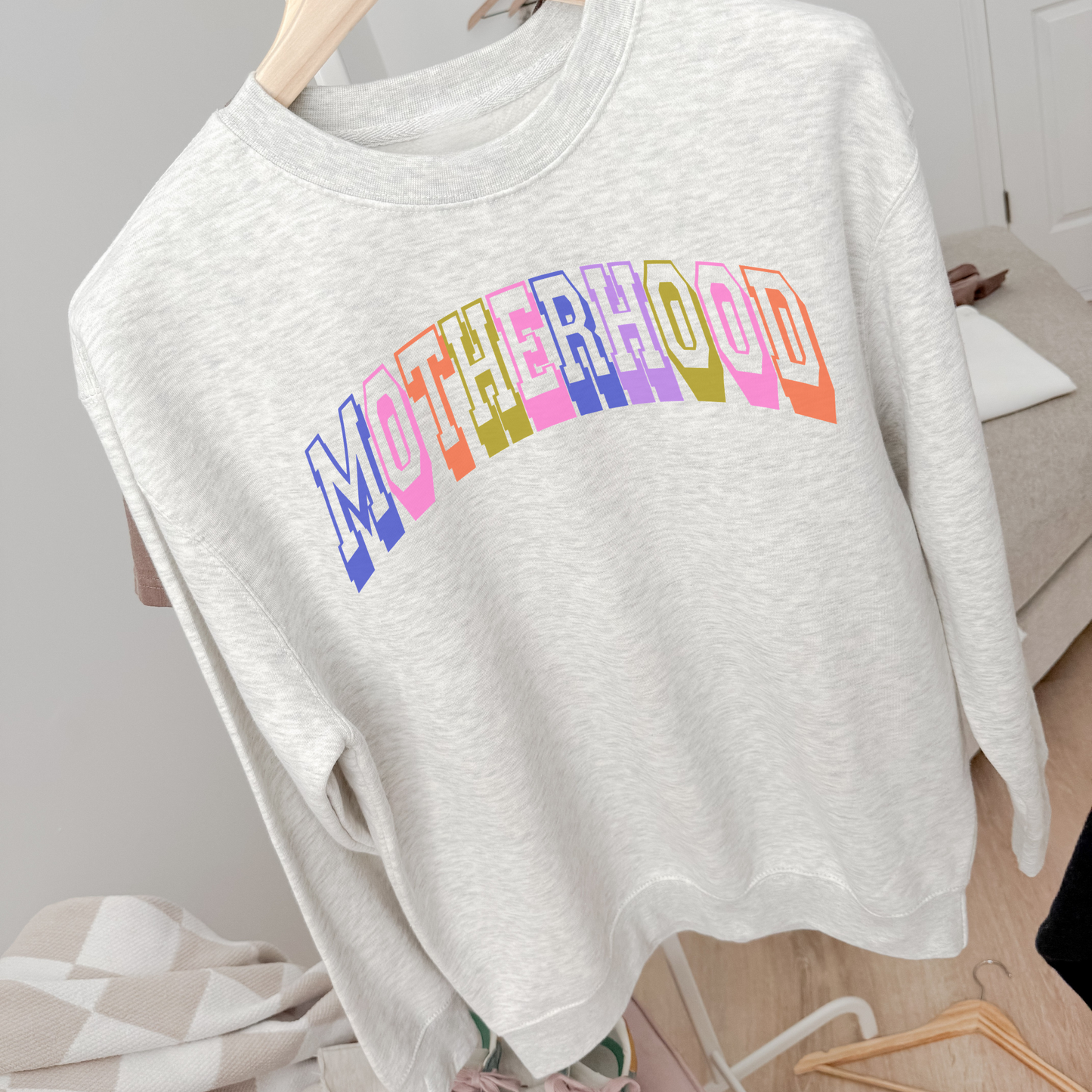 Ash grey motherhood graphic crewneck sweatshirt with colorful retro collegiate lettering hanging in cozy neutral room.