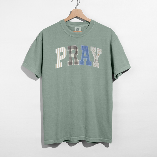 Green t-shirt with 'PRAY' printed on it, hanging on a wooden hanger against a white background.