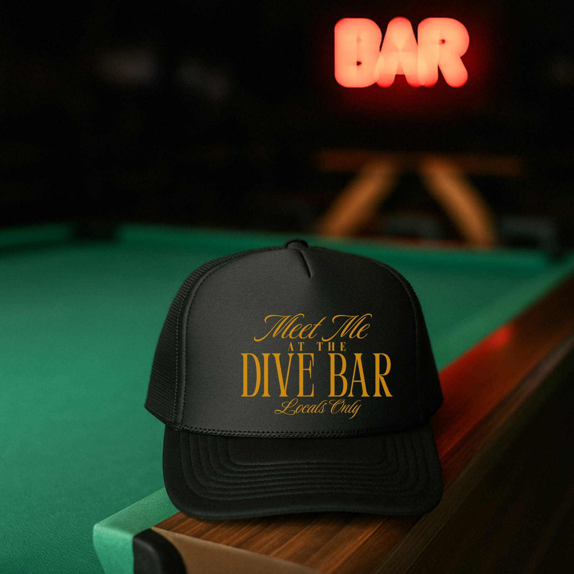 Black cap with 'Meet Me at the Dive Bar Locals Only' text on a pool table with neon 'BAR' sign in the background