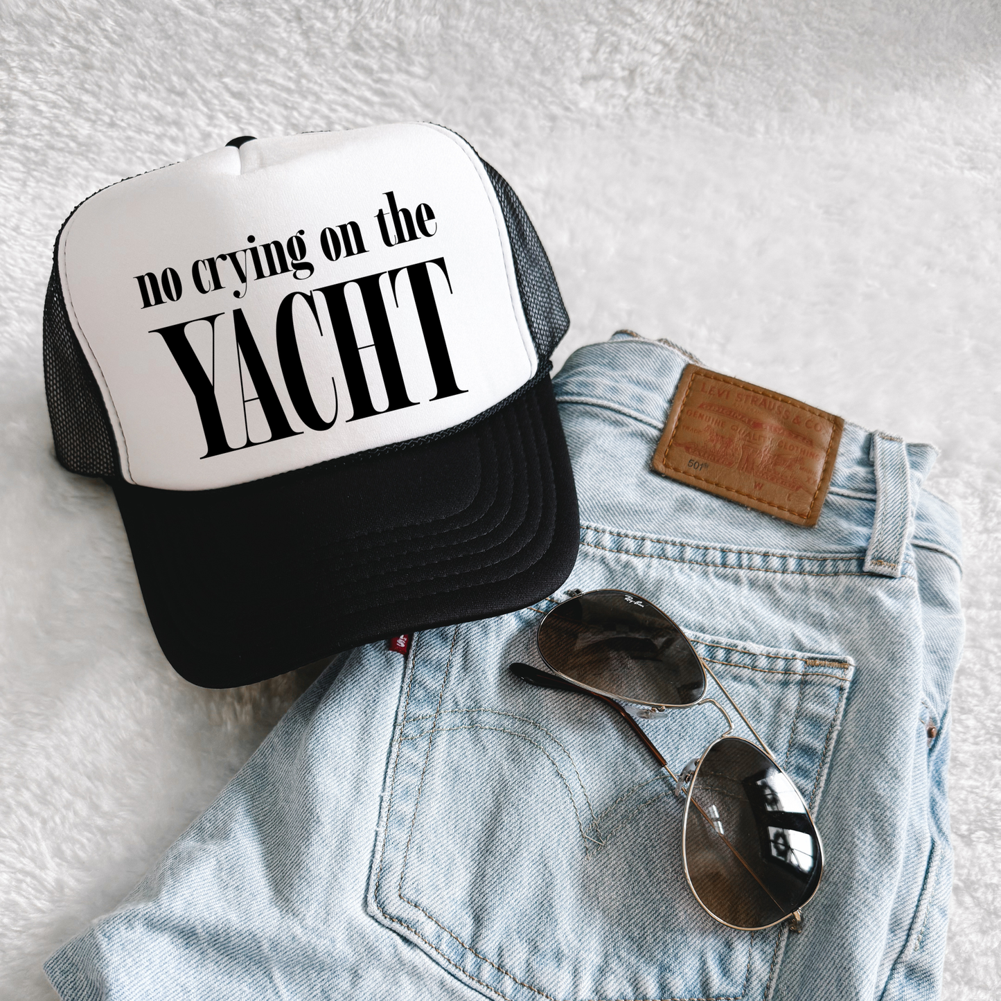 Black and white trucker hat with bold “No Crying on the Yacht” slogan displayed on folded denim jeans with sunglasses, lifestyle flat lay