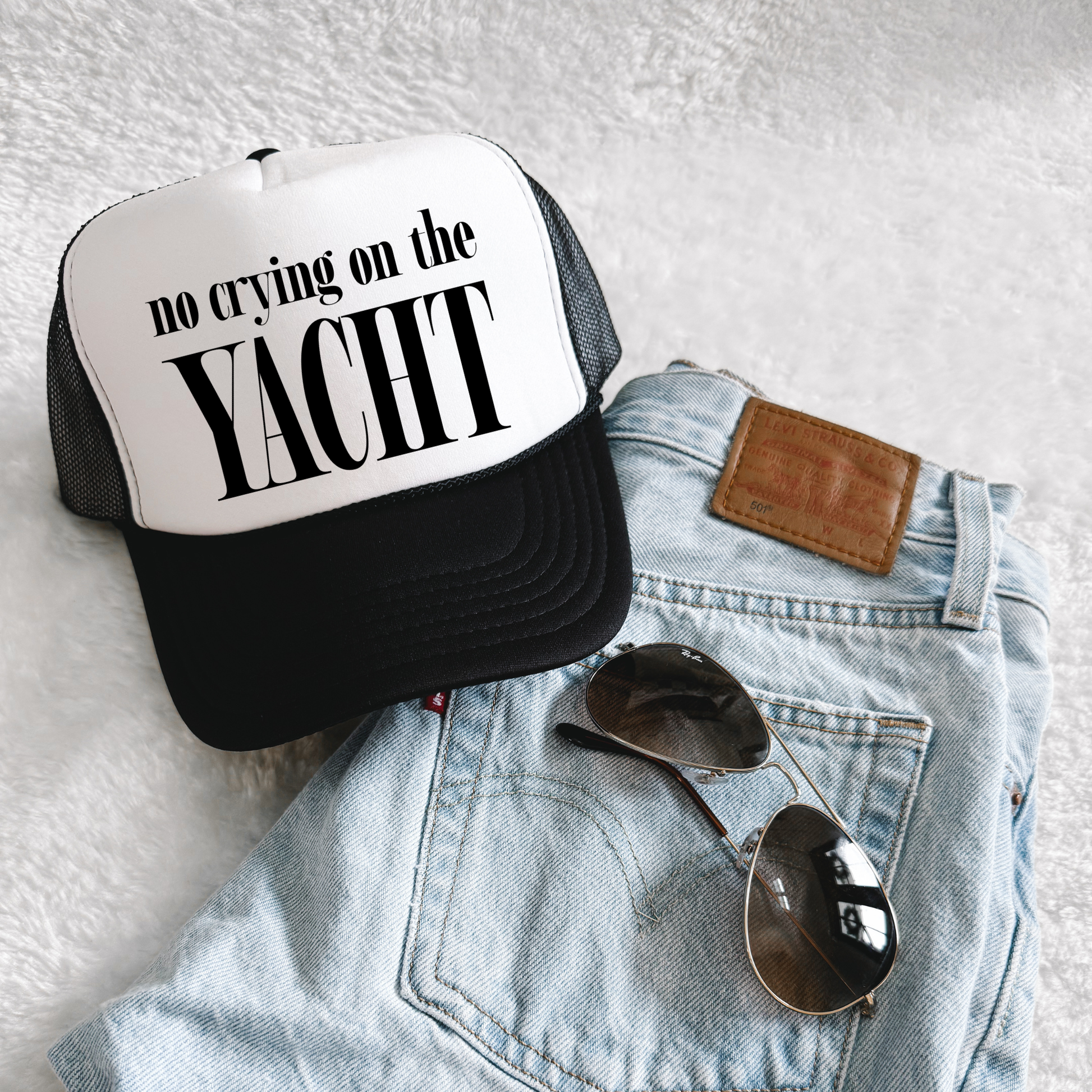 Black and white trucker hat with bold “No Crying on the Yacht” slogan displayed on folded denim jeans with sunglasses, lifestyle flat lay