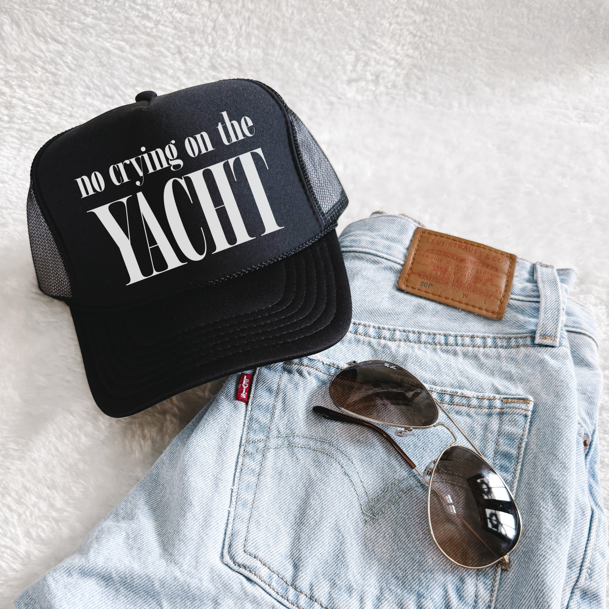 Black trucker hat with white “No Crying on the Yacht” text styled on light wash denim jeans with aviator sunglasses, flat lay product photo