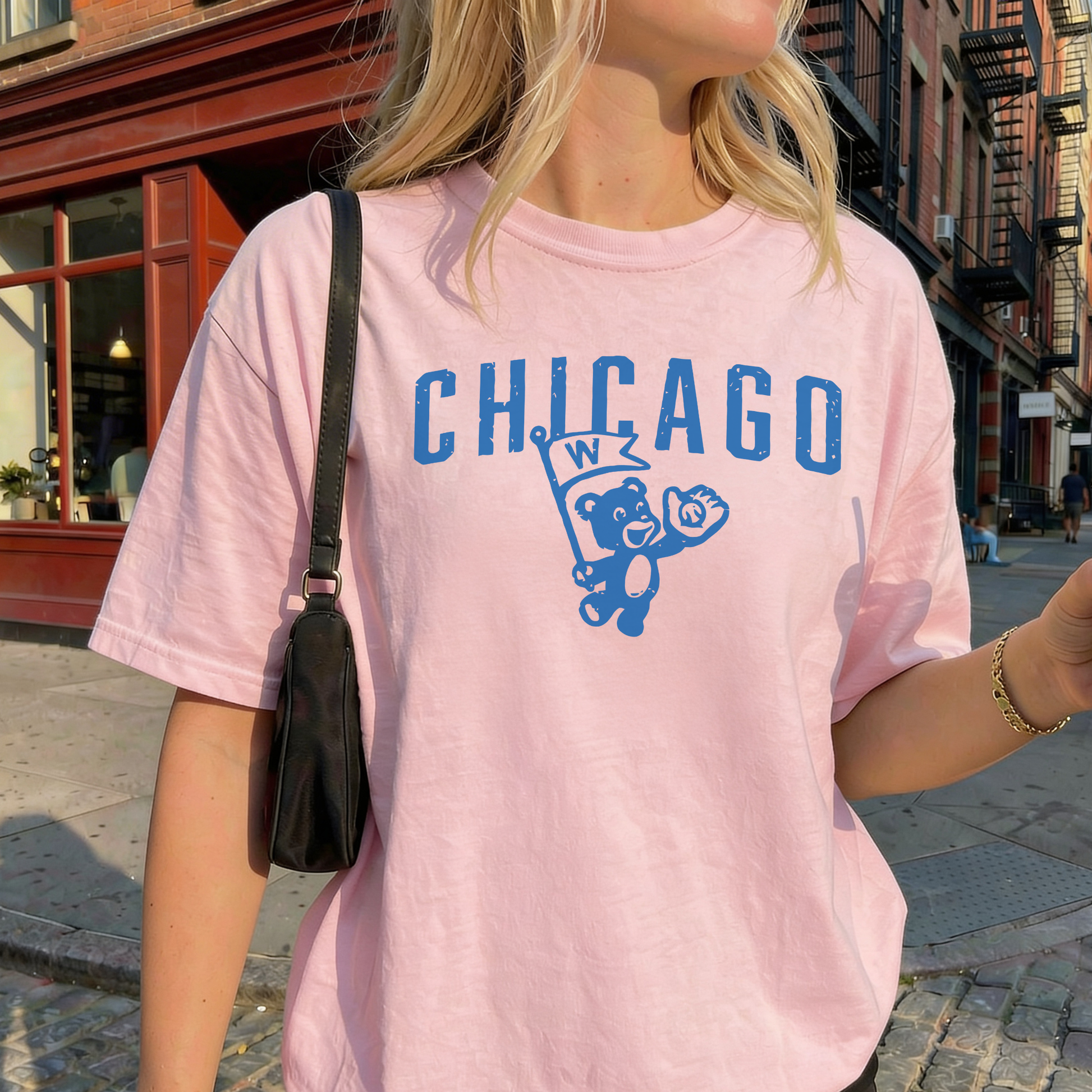Woman wearing blush pink Chicago retro cub graphic t-shirt styled with black shoulder bag on sunny urban sidewalk.