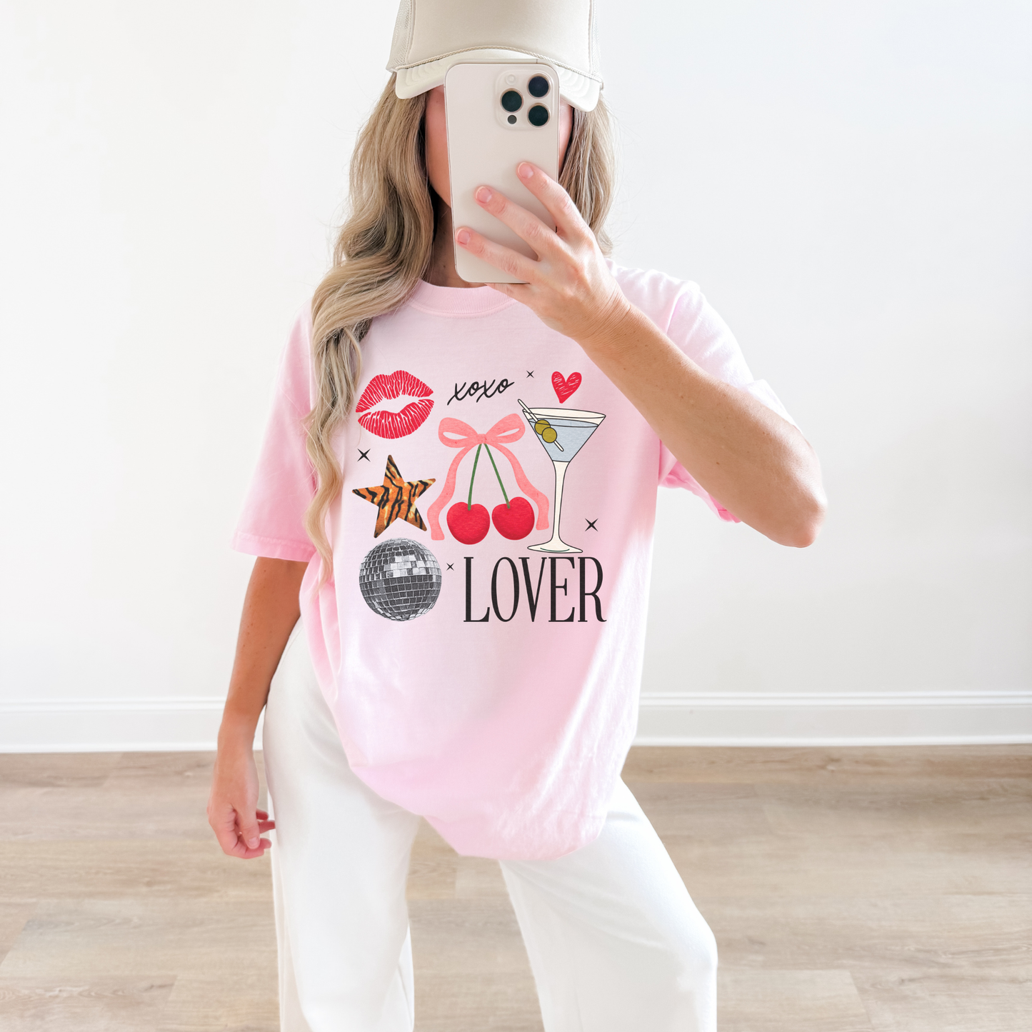 Woman wearing light pink Lover graphic tee with cherries, martini glass, and bow design, styled with white pants and baseball cap.