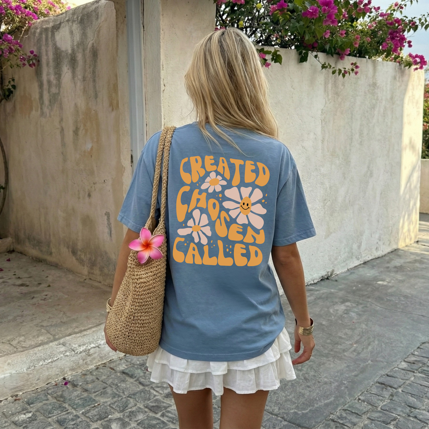Woman wearing a blue t-shirt with floral text design, standing outdoors near a stone wall with flowers.