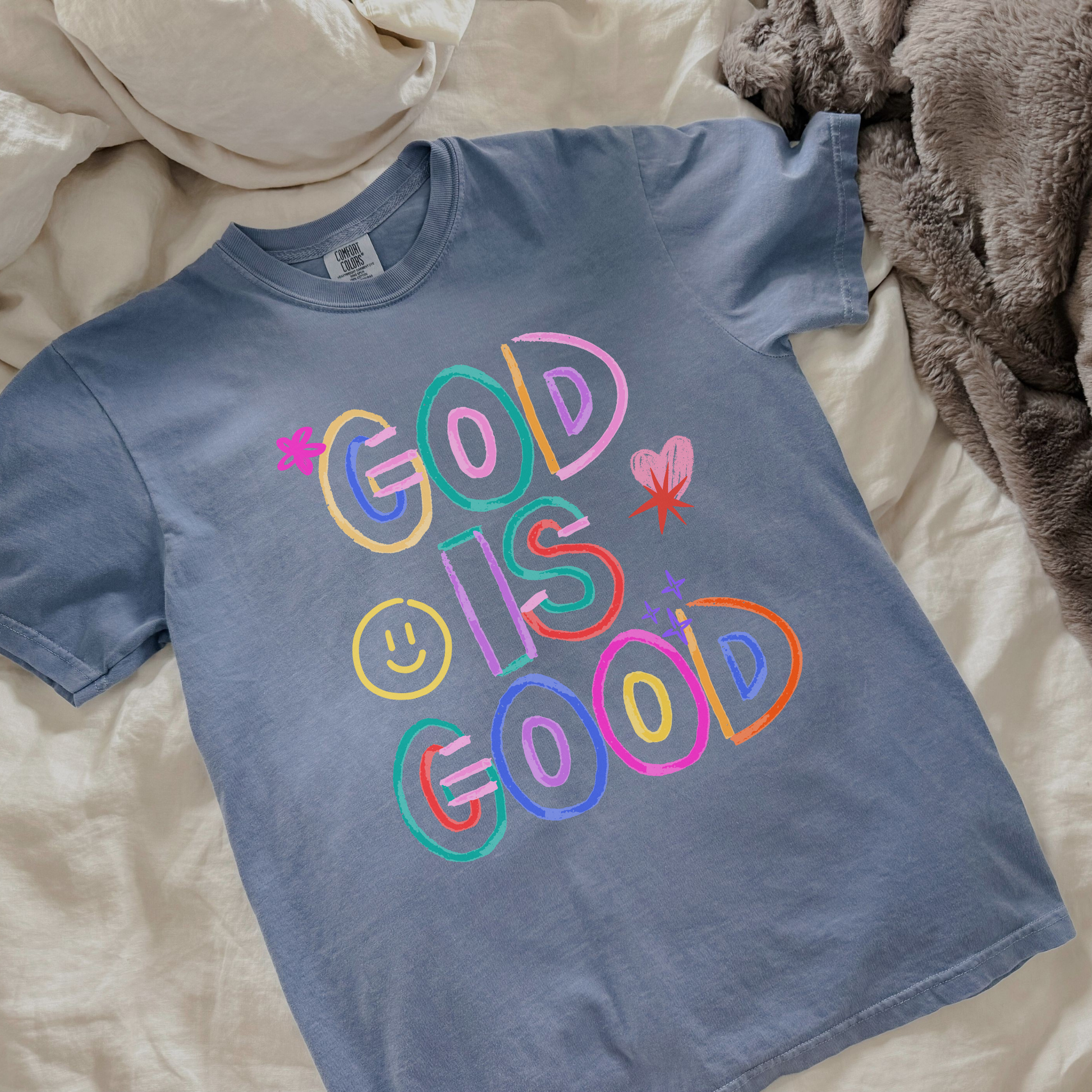 Blue t-shirt with colorful 'God is Good' text on a bed