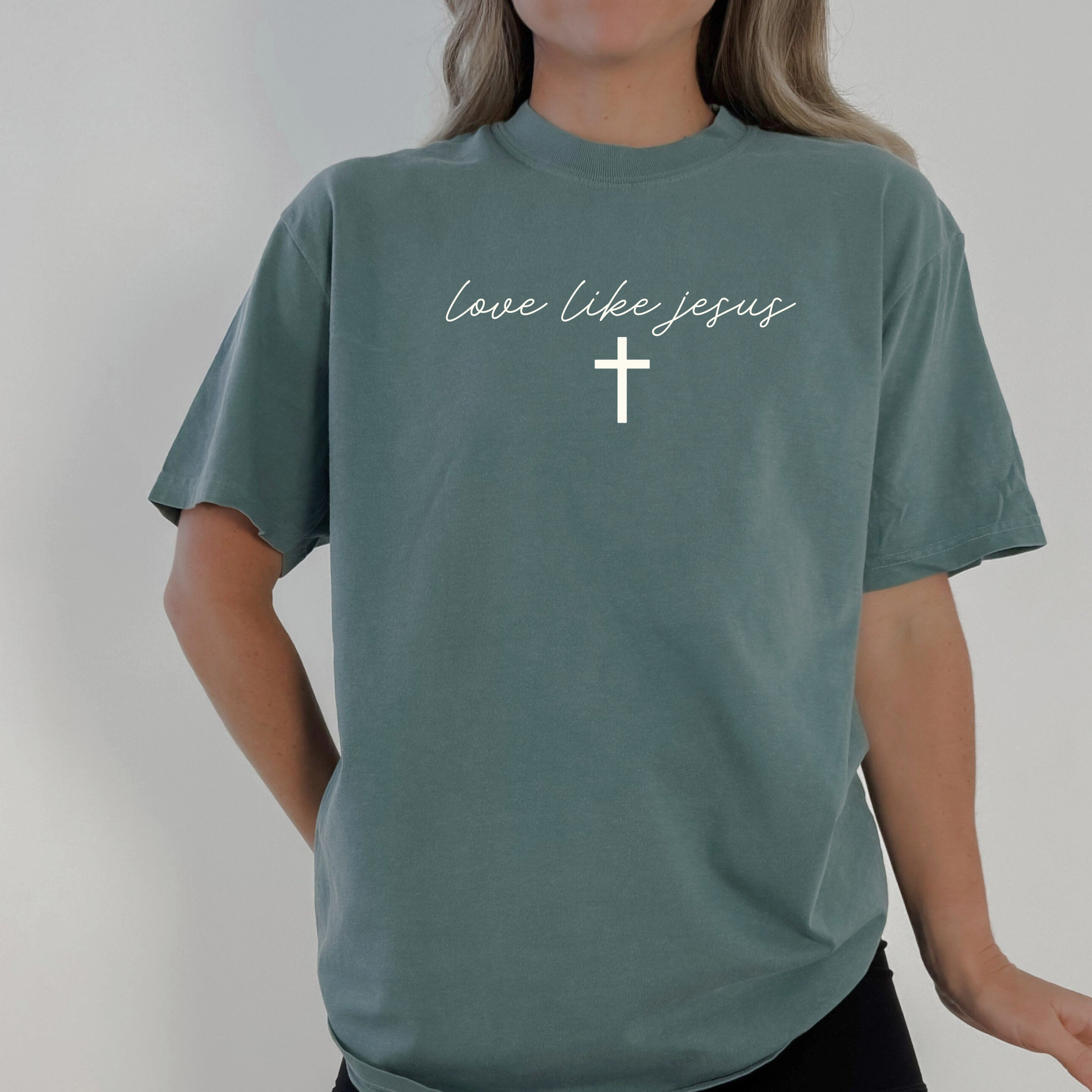 Woman wearing blue spruce Comfort Colors tee with “Love Like Jesus” script and cross; Christian inspirational graphic shirt.