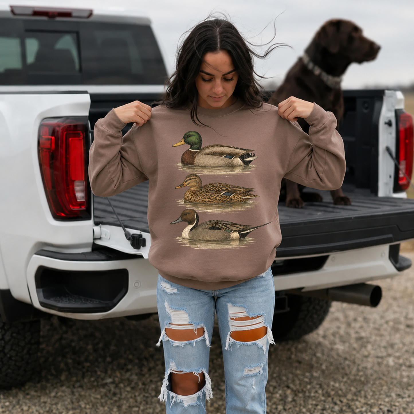 Woman wearing brown vintage waterfowl duck sweatshirt with distressed jeans beside pickup truck and hunting dog