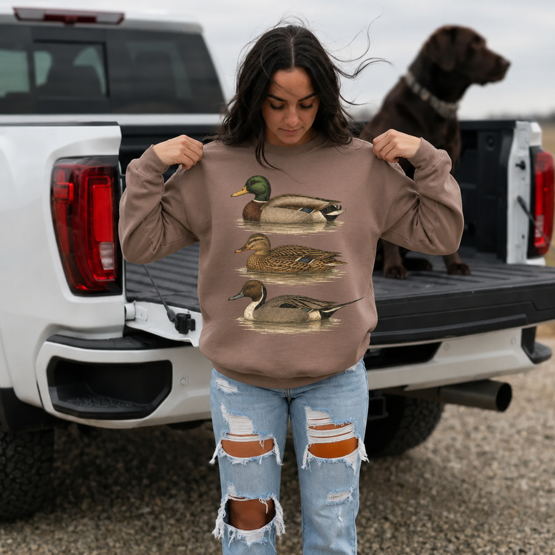 Woman wearing brown vintage waterfowl duck sweatshirt with distressed jeans beside pickup truck and hunting dog