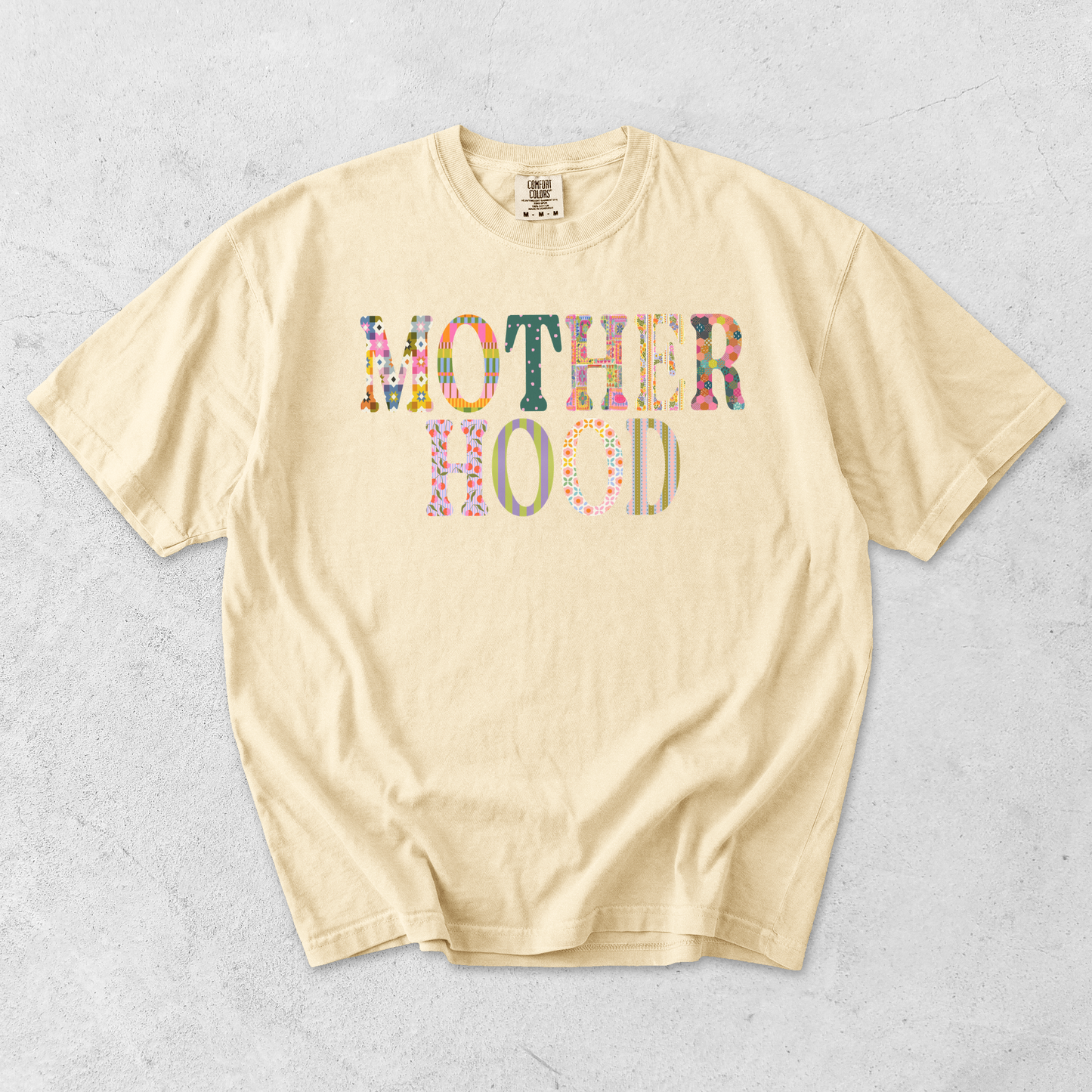 Butter yellow Comfort Colors motherhood graphic tee featuring colorful floral and striped patchwork lettering.