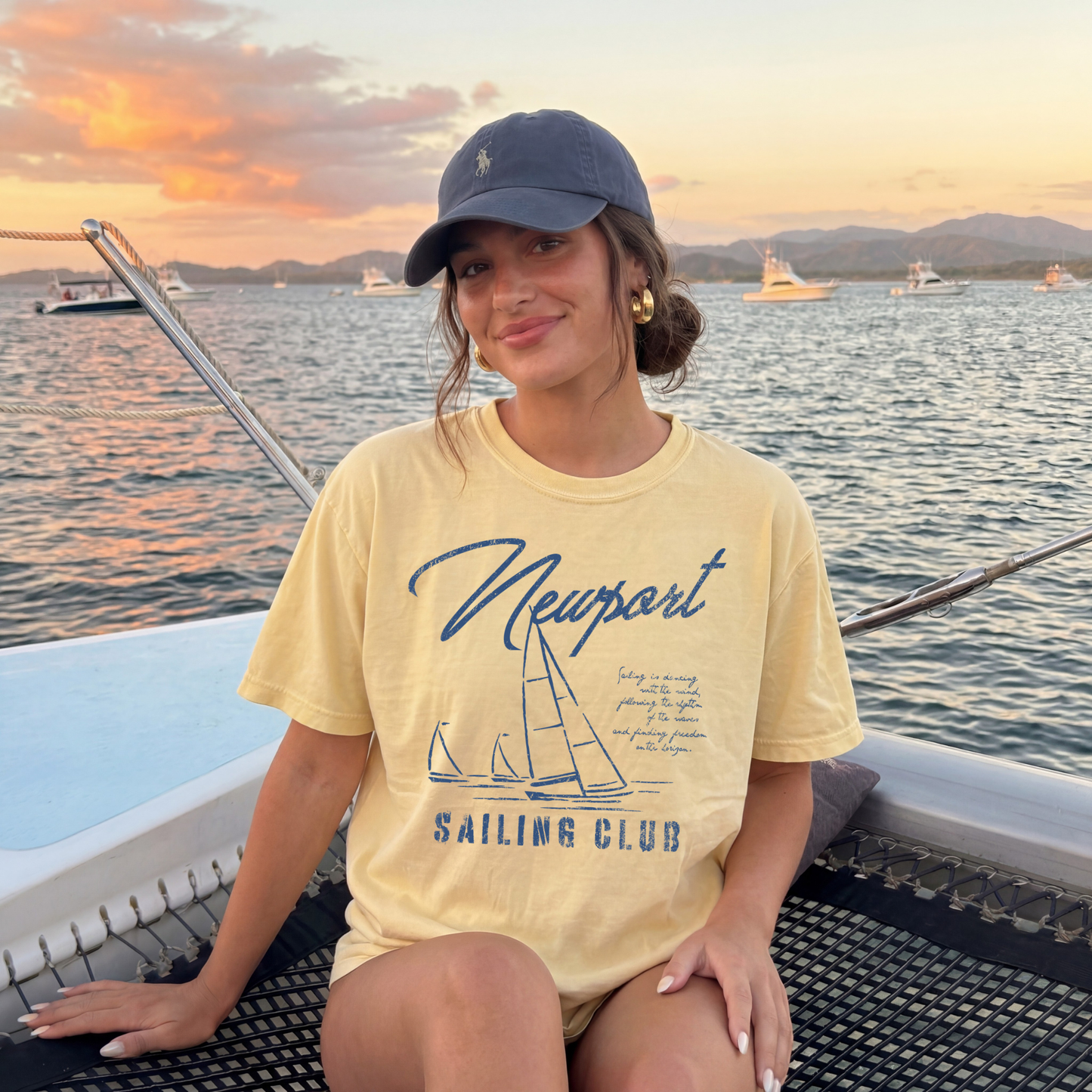 woman wearing butter yellow sailing club graphic tee on boat at sunset coastal summer outfit