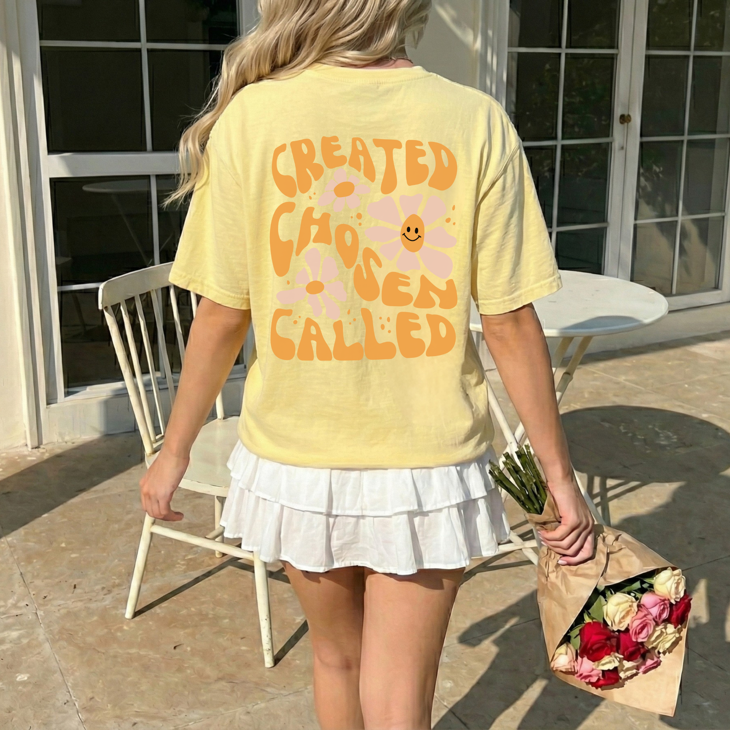 Person wearing a yellow t-shirt with text and flowers, holding flowers outdoors.