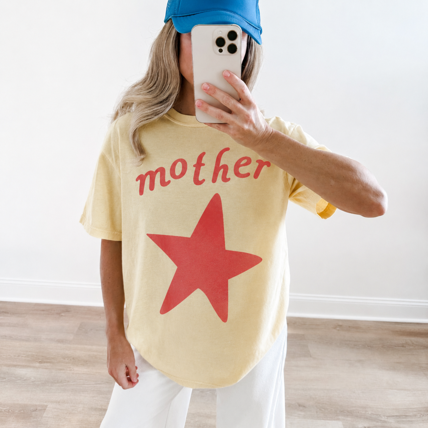 Woman wearing a butter yellow Mother graphic tee featuring a vintage coral star and retro lettering with white wide-leg pants and a blue trucker hat.