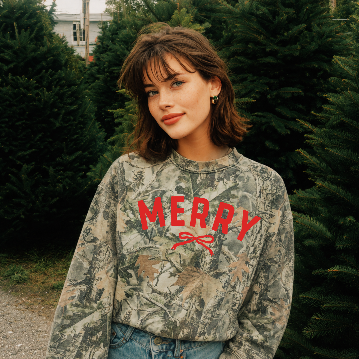 Woman wearing a premium oversized camo Merry Christmas crewneck with red bow graphic at a Christmas tree farm.
