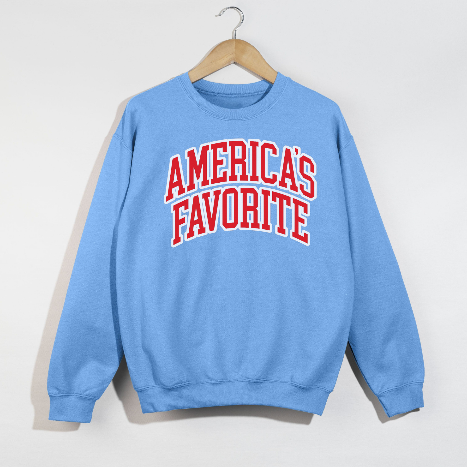 Carolina blue patriotic crewneck sweatshirt featuring bold red and white “America’s Favorite” collegiate lettering hanging on wooden hanger.