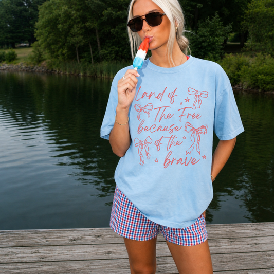 Woman wearing a chambray Comfort Colors 1717 graphic tee with pink “Land of the Free Because of the Brave” patriotic bow design while holding a red, white, and blue popsicle on a lakeside dock.
