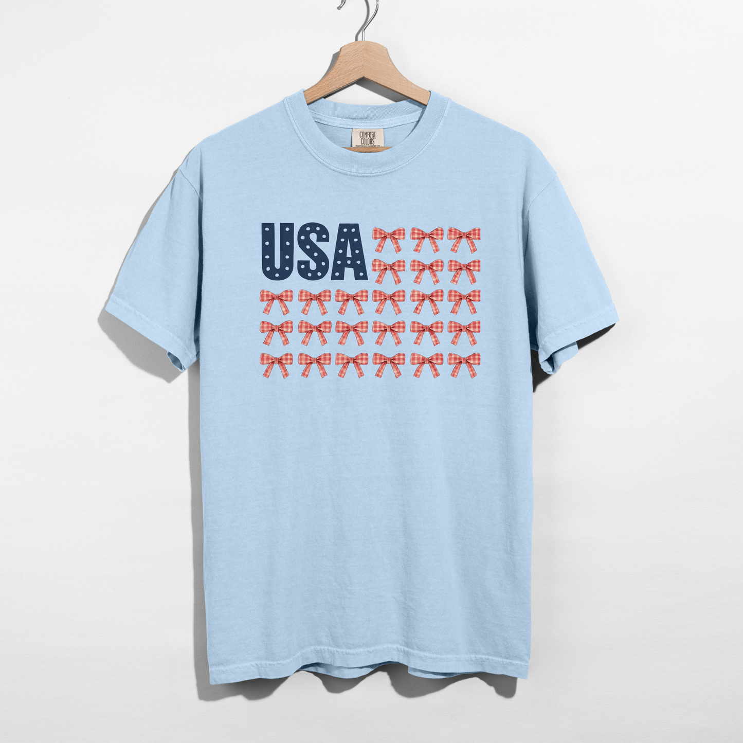 Light blue t-shirt with 'USA' and bow design on a hanger against a white background