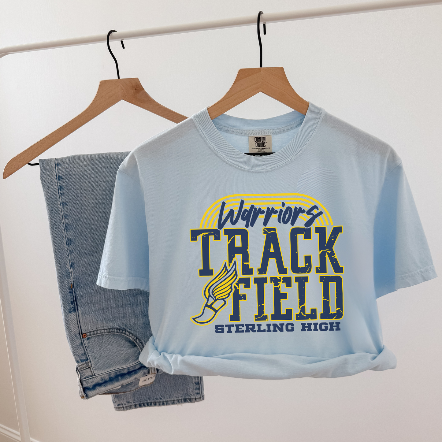 Light blue t-shirt with 'Warriors Track Field Sterling High' design on a hanger against a white background.