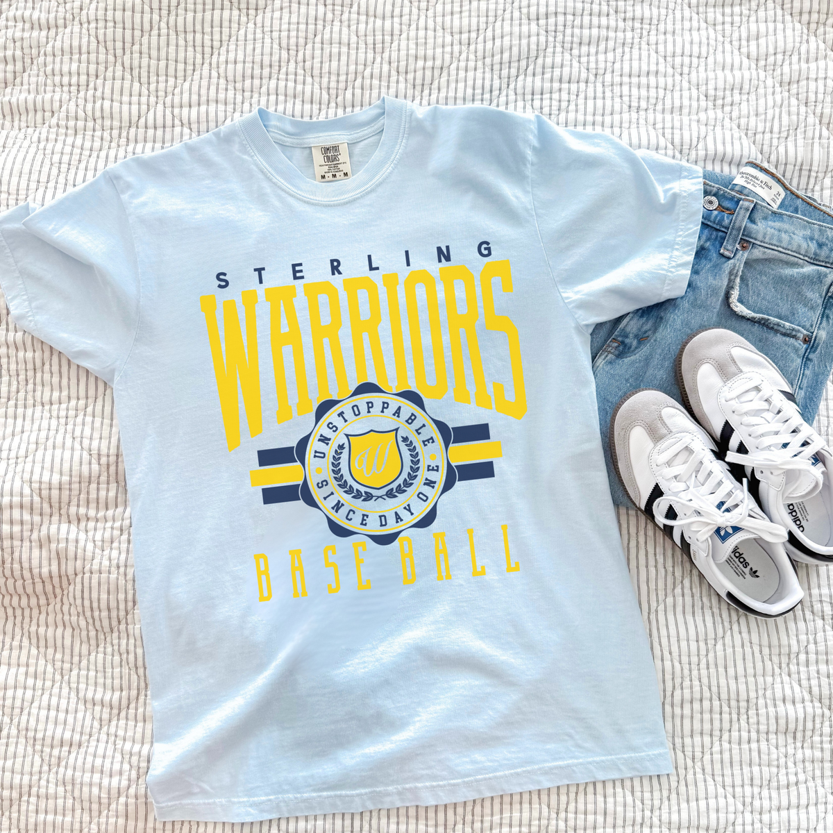 Light blue t-shirt with 'Sterling Warriors Baseball' design on a textured surface with sneakers and jeans.