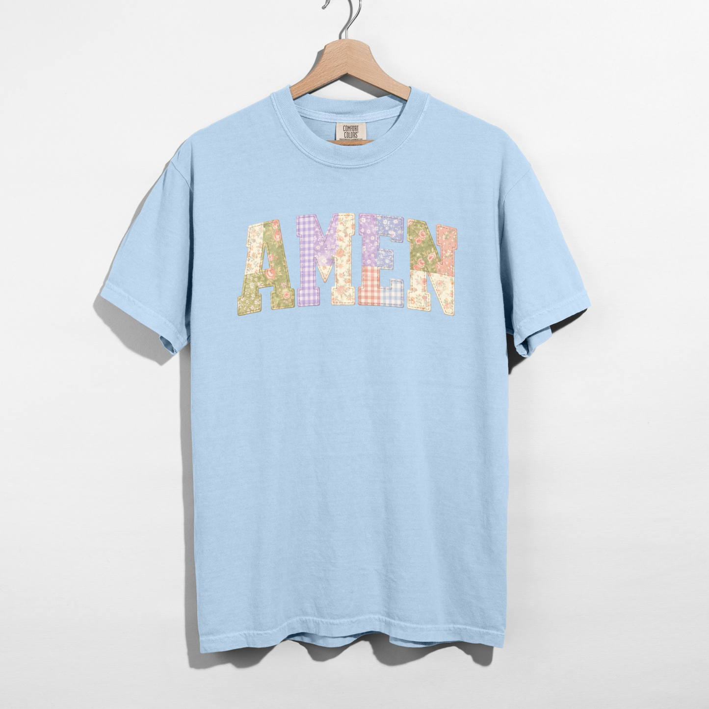 Light blue t-shirt with 'AMEN' printed in colorful letters on a hanger against a white background