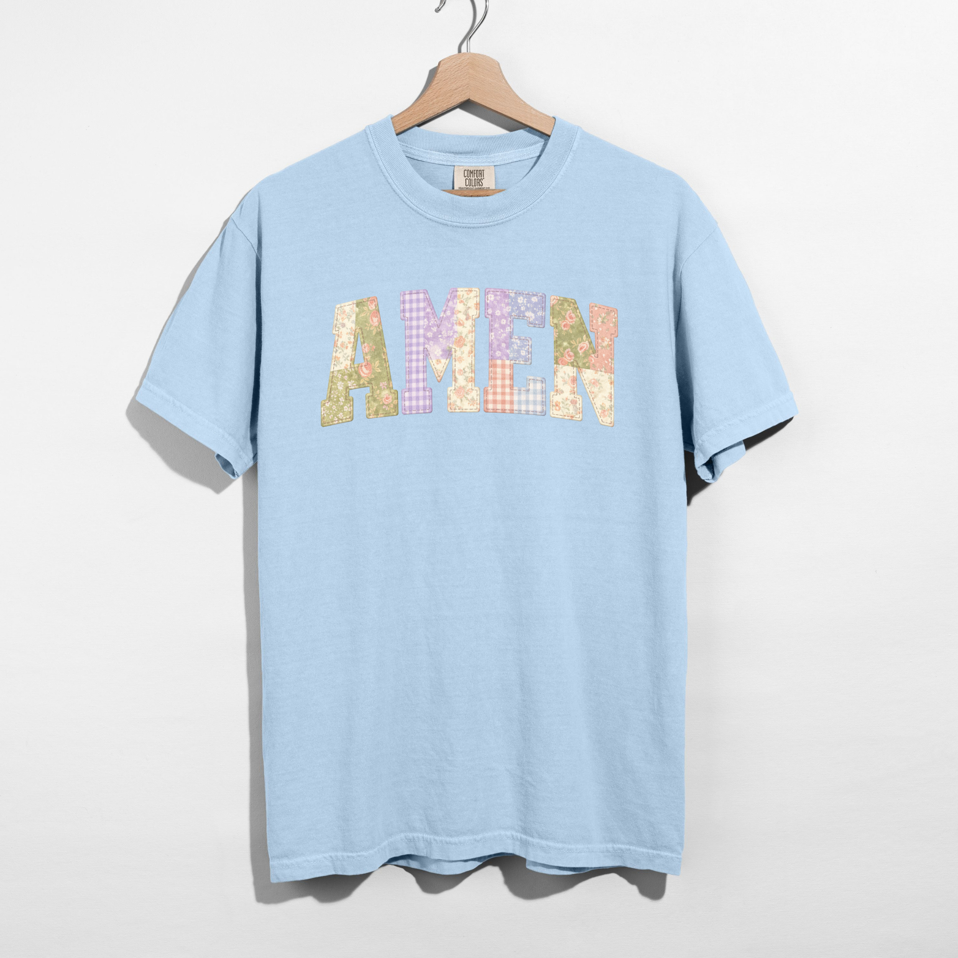 Light blue t-shirt with 'AMEN' printed in colorful letters on a hanger against a white background