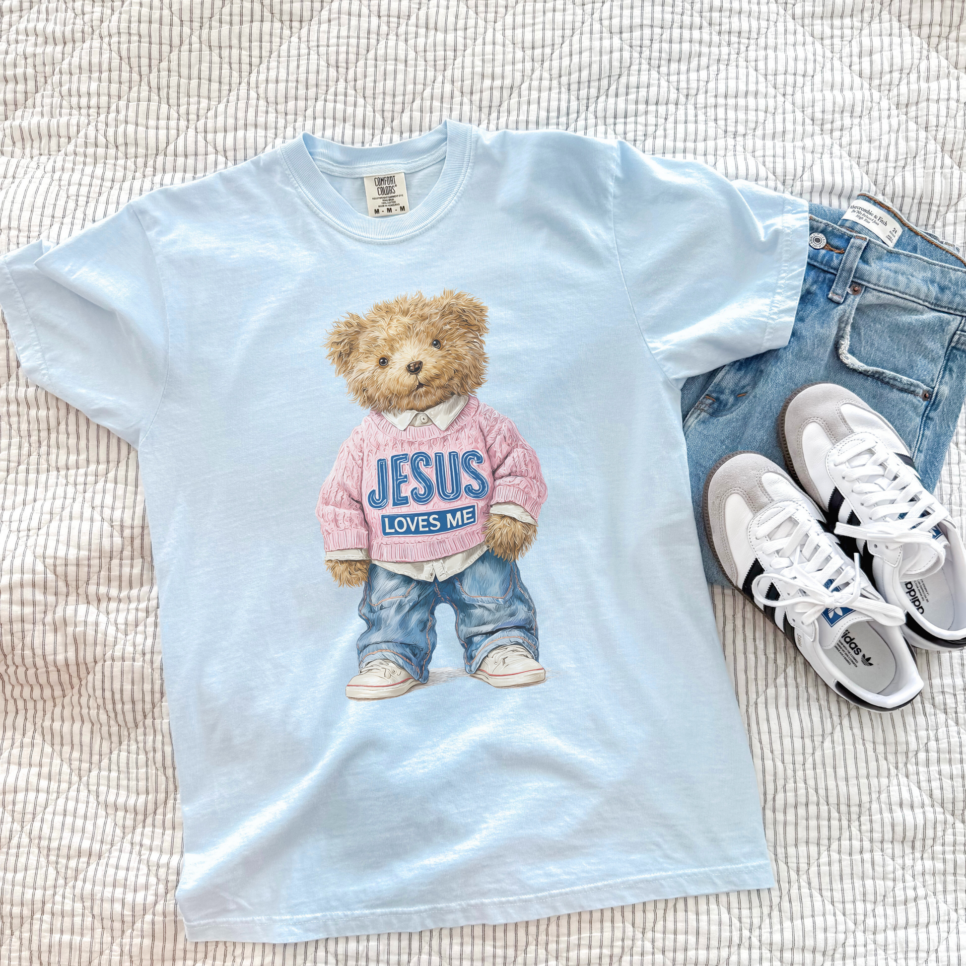 Light blue t-shirt with teddy bear graphic and 'Jesus loves me' text, paired with jeans and sneakers on a textured surface.