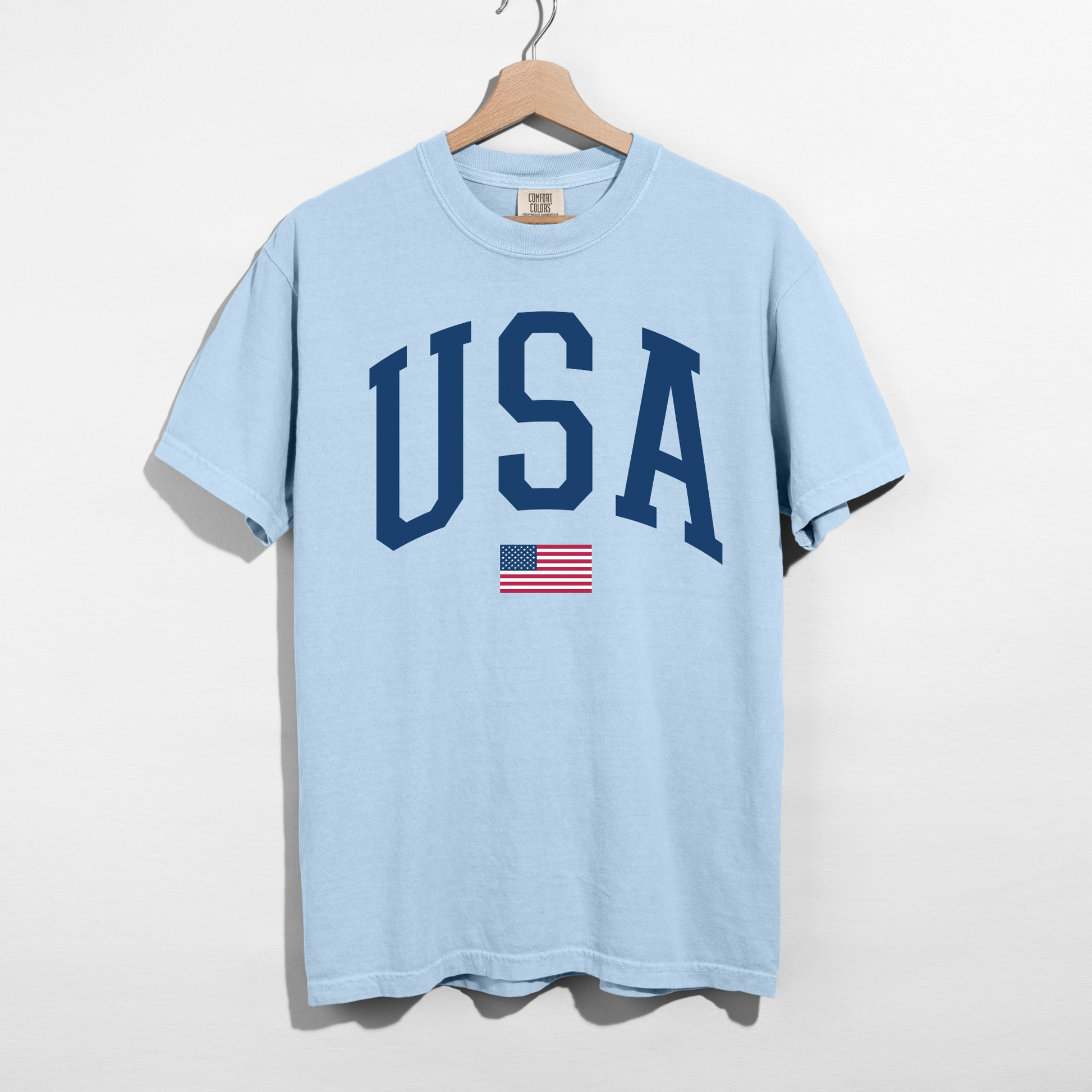 Light blue USA varsity graphic tee with American flag detail hanging on wooden hanger