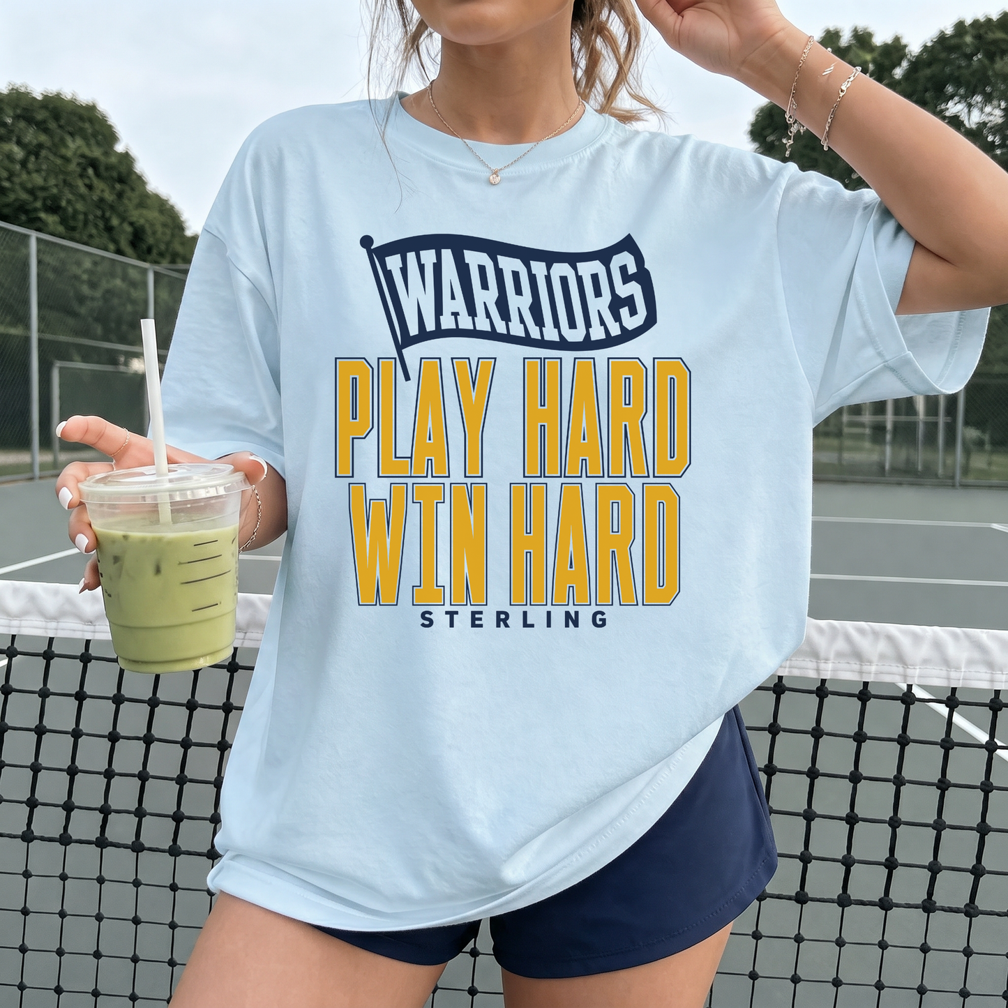 Chambray blue Comfort Colors Sterling Warriors Play Hard Win Hard graphic t-shirt styled on model at outdoor tennis courts with navy athletic shorts.