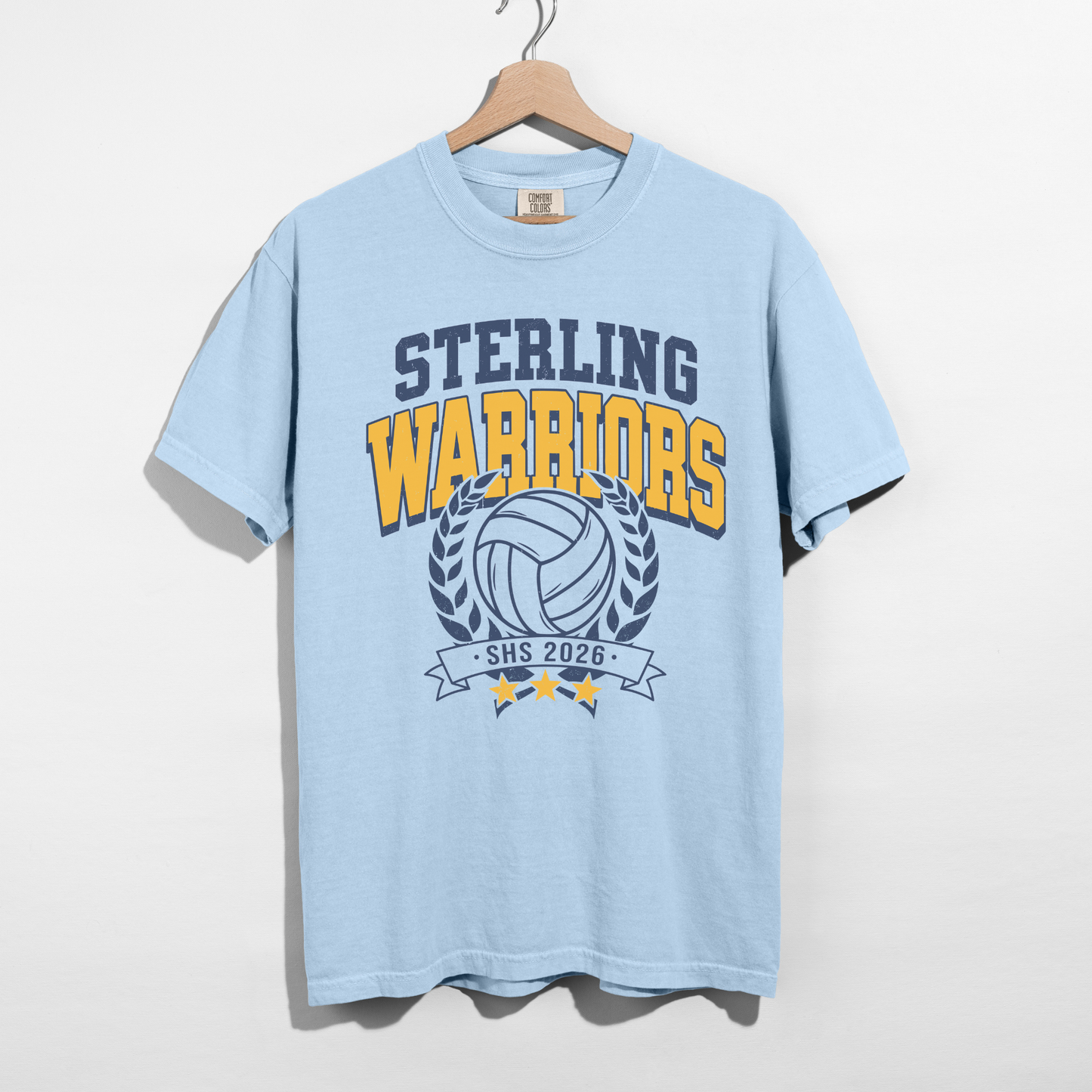 Chambray blue Sterling Warriors Volleyball Comfort Colors graphic tee hanging on wooden hanger featuring navy and gold volleyball school spirit design.
